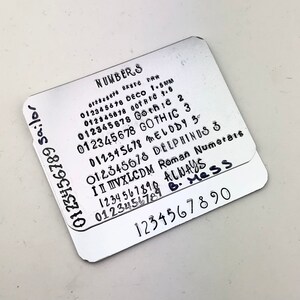 May include: Two silver metal plates with different font styles of numbers 0-9. The top plate has the words "NUMBERS" and "ALWAYS B. Mess" written on it. The bottom plate has the numbers 1234567890 written on it.