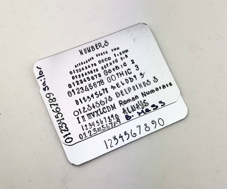 May include: Two silver metal plates with different font styles of numbers 0-9. The top plate has the words "NUMBERS" and "ALWAYS B. Mess" written on it. The bottom plate has the numbers 1234567890 written on it.