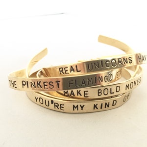 May include: A set of four gold-toned metal bracelets with different inspirational phrases engraved on them. The phrases are "Real Unicorns Have The Pinkest Flamingos", "Make Bold Moves", and "You're My Kind Of".