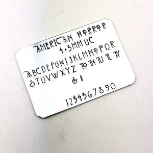 May include: A silver metal stamp with the text "AMERICAN HORROR 4.5MM UC" and the alphabet in uppercase letters, followed by the ampersand and number sign, and the numbers 1 through 90.
