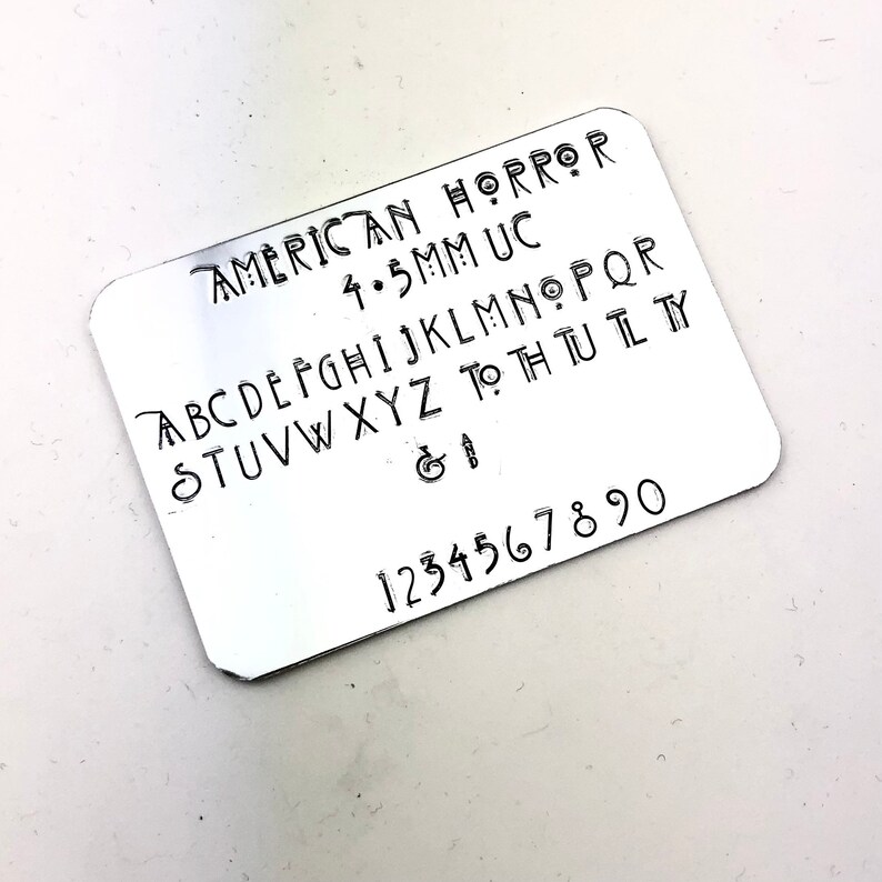 May include: A silver metal stamp with the text "AMERICAN HORROR 4.5MM UC" and the alphabet in uppercase letters, followed by the ampersand and number sign, and the numbers 1 through 90.