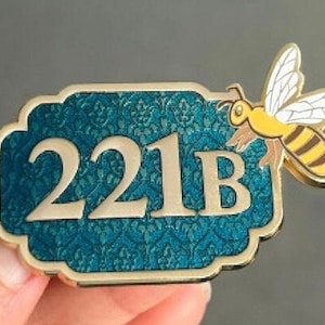 May include: A gold-colored metal sign with a teal blue background and a white floral pattern. The sign reads "221B" in gold lettering. A gold-colored bee with black stripes and white wings is attached to the sign.