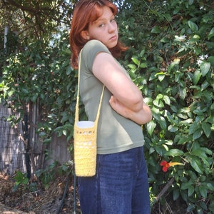 May include: A person wearing a green shirt and blue jeans is carrying a yellow crocheted water bottle holder.