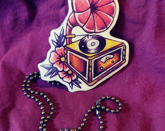 May include: A pendant featuring a vintage-style gramophone design with a red horn, a record, and floral accents. The pendant is attached to a dark metal ball chain, creating a unique accessory.