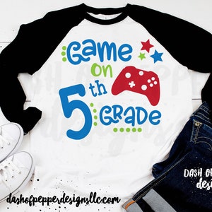 May include: A white t-shirt with a black and red baseball sleeve design. The shirt has a blue number 5 with green dots, the text "Game On" in green and red, and a red video game controller graphic.