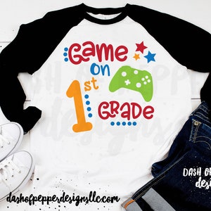 Könnte beinhalten: Ein weißes und schwarzes Baseball-T-Shirt mit einem bunten Grafikdesign, auf dem "Game on 1st Grade" mit einem grünen Videospiel-Controller steht. Das Design enthält die Farben Rot, Orange, Gelb, Blau und Grün.