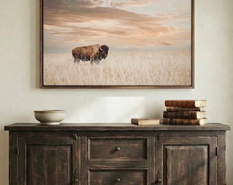 May include: A rectangular framed wall art print featuring a brown bison standing in a vast field of dry golden grass under a soft, cloudy sunset sky. The artwork is displayed above a dark brown wooden sideboard with three drawers and two cabinets.