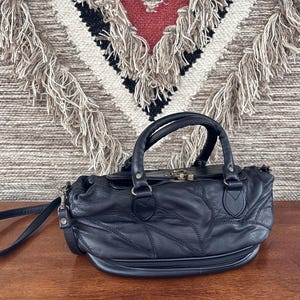 May include: Black leather handbag with a top handle and a detachable shoulder strap. The bag has a patchwork design and a zipper closure. The background features a woven textile with fringe.