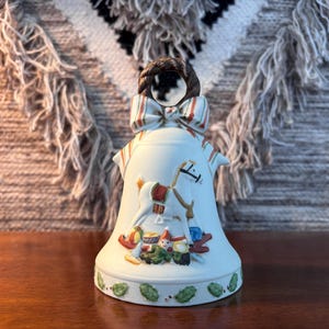May include: A white ceramic bell with a decorative design featuring a rocking horse and toy soldiers. The bell has a striped bow and a brown hair-like element at the top. The base is adorned with green leaves.