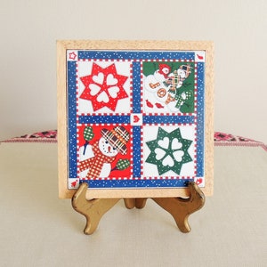May include: A square decorative tile in a light wood frame featuring a four-panel grid design. The panels display red and green star patterns with heart cutouts and illustrations of snowmen wearing plaid hats and scarves on a blue background.