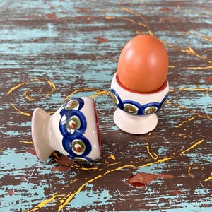 May include: Two ceramic egg cups featuring a white base with a hand-painted pattern of blue circles and green centers. One egg cup is positioned upright holding a single brown egg, while the other egg cup is lying on its side next to it.