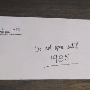Puede incluir: Un sobre blanco con el texto "Lou's Cafe, Hill and Main, Hill Valley, California" y el texto escrito a mano "No abrir hasta 1985".