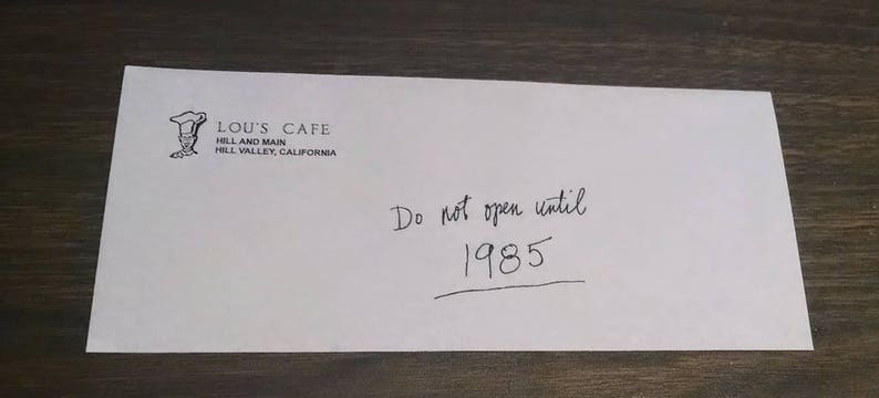Puede incluir: Un sobre blanco con el texto "Lou's Cafe, Hill and Main, Hill Valley, California" y el texto escrito a mano "No abrir hasta 1985".