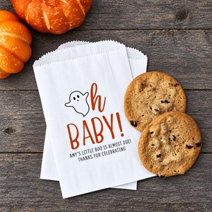 May include: A white paper treat bag featuring a black ghost illustration, the orange text "oh BABY!", and the black text "AMY'S LITTLE BOO IS ALMOST DUE! THANKS FOR CELEBRATING". Two round chocolate chip cookies are placed next to the paper bag.