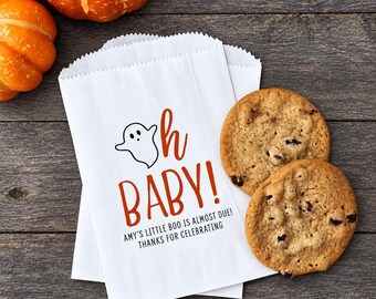 May include: A white paper treat bag featuring a black ghost illustration, the orange text "oh BABY!", and the black text "AMY'S LITTLE BOO IS ALMOST DUE! THANKS FOR CELEBRATING". Two round chocolate chip cookies are placed next to the paper bag.
