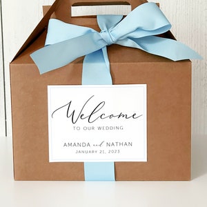 May include: A brown cardboard gift box with a blue ribbon and a white label that says "Welcome to our wedding Amanda and Nathan January 21, 2023".