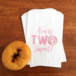 May include: A golden-brown donut sits next to three white paper treat bags. The bags have a pink text that reads "Ava is TWO Sweet!" with a donut graphic. The bags are on a wooden surface.