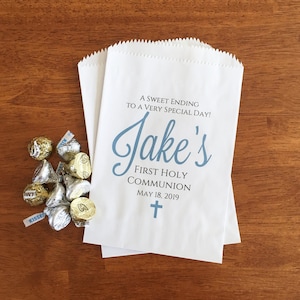 May include: White paper favour bags with a blue cross and the text "A sweet ending to a very special day! Jake's First Holy Communion May 18, 2019".