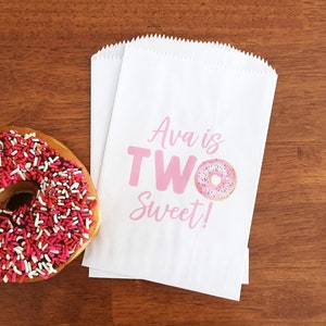 May include: Two white paper treat bags with pink text that reads "Ava is TWO Sweet!" and a donut graphic. A glazed donut with pink and red sprinkles sits to the left on a wooden surface.