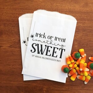 May include: Two white paper bags with serrated top edges featuring black text that reads trick or treat something SWEET 10th ANNUAL HALLOWEEN BLOCK PARTY and a spider graphic. Beside the bags are several orange, yellow, and white candy corns.