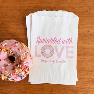 May include: White paper favor bags with pink text that reads "Sprinkled with LOVE" and a pink donut with sprinkles. The text below the donut reads "Emily's Baby Sprinkle".