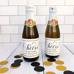 May include: Two small glass bottles with gold and black labels that say "Cheers! To 50 Years!" and "Sheila's Birthday Dinner 8.2.20".