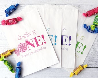 May include: Four white paper bags with colorful text that reads "Amelia is ONE!" and "Thanks for celebrating". The bags are filled with colorful candy.