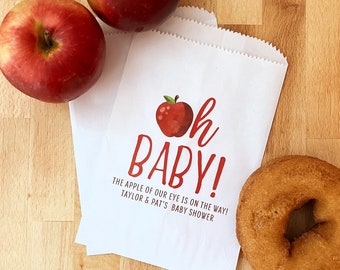 May include: White paper bag with red apple graphic and the text "Oh Baby! The apple of our eye is on the way! Taylor & Pat's Baby Shower".