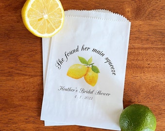 May include: White favor bag with a lemon design and the text "She found her main squeeze" and "Heather's Bridal Shower 4.1.2022".