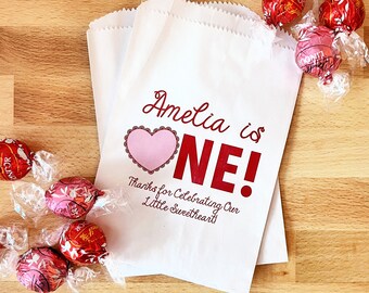 May include: White favor bags with red and pink text that says "Amelia is ONE!" and "Thanks for Celebrating Our Little Sweetheart". The bags are filled with red and pink chocolate candies.