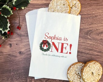 May include: Two white paper treat bags with a red and green wreath design and the text "Sophia is ONE!" and "Thanks for celebrating with us!" Two round crackers are visible. The bags are on a wooden surface.