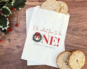 May include: White paper gift bag with a red and green Christmas wreath design. The bag has the text "Oh, what fun to be ONE! Sophia's 1st Birthday" printed on it.