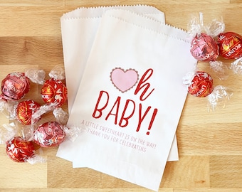 Könnte beinhalten: Weiße Papiergeschenktüten mit rosa und roten Herzen und dem Text "Oh Baby! A little sweetheart is on the way! Thank you for celebrating." Die Taschen sind mit roten Schokoladenbonbons gefüllt.