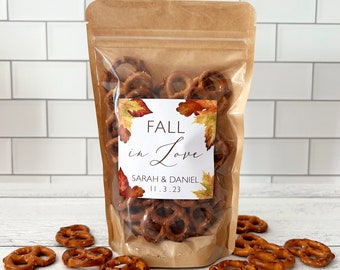 May include: A clear plastic bag filled with pretzel bites, with a brown paper label that reads "Fall in Love" with the names "Sarah & Daniel" and the date "11.3.23". The bag is sitting on a white tiled surface with a few pretzel bites scattered around it.
