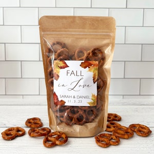May include: A clear plastic bag filled with pretzel bites, with a brown paper label that reads "Fall in Love" with the names "Sarah & Daniel" and the date "11.3.23". The bag is sitting on a white tiled surface with a few pretzel bites scattered around it.