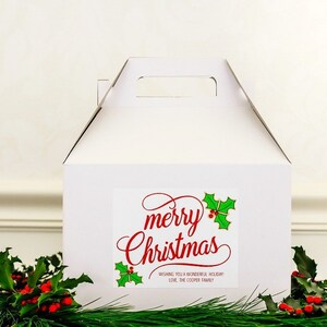 May include: White gift box with a handle. The box has a sticker with the text "merry Christmas" in red cursive lettering with green holly leaves and red berries. The sticker also says "Wishing you a wonderful holiday Love, The Cooper Family."