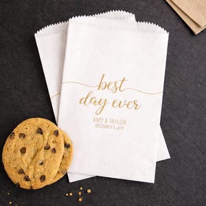 May include: White paper treat bags with a scalloped top, printed with "best day ever" in gold script, along with a date and names. Two chocolate chip cookies are next to the bags. A brown napkin is in the background.