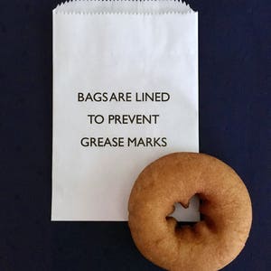 May include: A white paper bag with the text "BAGS ARE LINED TO PREVENT GREASE MARKS" printed on it. A brown donut is sitting next to the bag.