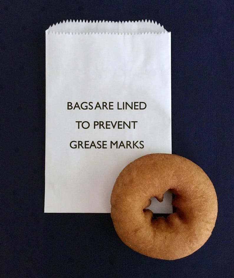 May include: A white paper bag with the text "BAGS ARE LINED TO PREVENT GREASE MARKS" printed on it. A brown donut is sitting next to the bag.