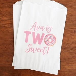 May include: Two white paper treat bags with a pink graphic that reads "Ava is TWO Sweet!" and a donut illustration. The bags are on a brown wooden surface.
