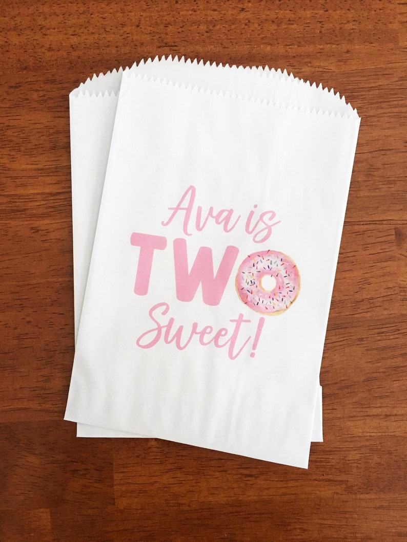 May include: Two white paper treat bags with a pink graphic that reads "Ava is TWO Sweet!" and a donut illustration. The bags are on a brown wooden surface.