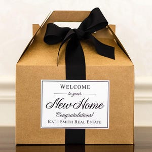 May include: A brown gift box with a black ribbon and bow. The box has a white label with the text "WELCOME to your New Home Congratulations! KATE SMITH REAL ESTATE".