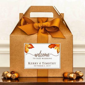 May include: A brown cardboard box with a gold ribbon and a white label. The label has a watercolor design of fall leaves and the text "welcome to our wedding Kerry & Timothy October 5, 2019".
