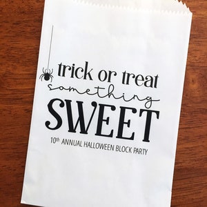 May include: A white paper bag with a serrated top edge featuring black printed text that reads trick or treat something SWEET 10th ANNUAL HALLOWEEN BLOCK PARTY and a small black spider graphic hanging from a thin vertical line on the left side.