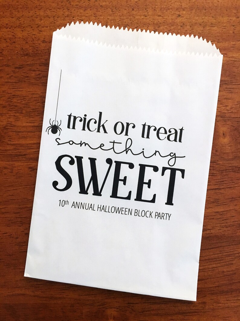 May include: A white paper bag with a serrated top edge featuring black printed text that reads trick or treat something SWEET 10th ANNUAL HALLOWEEN BLOCK PARTY and a small black spider graphic hanging from a thin vertical line on the left side.