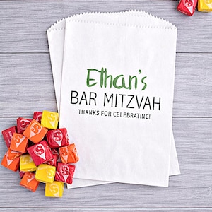 May include: White paper favor bags with the text "Ethan's BAR MITZVAH" in green and black. Colorful candies in red, orange, and yellow are scattered around the bags on a gray wooden surface.