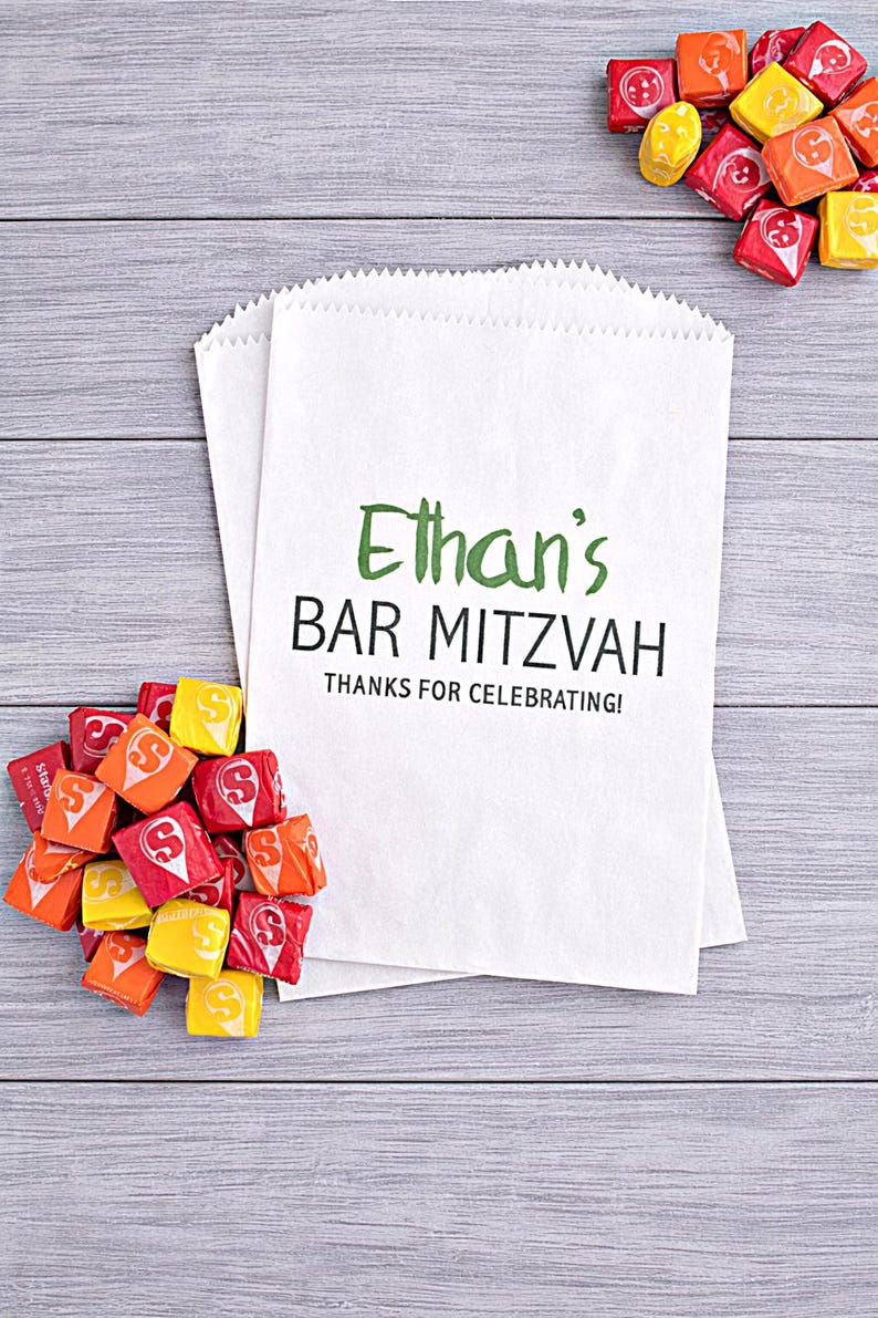 May include: White paper favor bags with the text "Ethan's BAR MITZVAH" in green and black. Colorful candies in red, orange, and yellow are scattered around the bags on a gray wooden surface.
