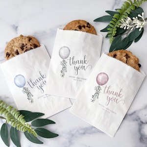 May include: Three white paper bags with cookies inside. Each bag has a different colored balloon with a thank you message and the text "Kristin's Baby Shower" and the date "May 4, 2024".