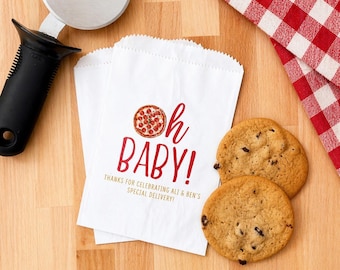 May include: Two white paper bags with the text "Oh Baby!" in red, featuring a pizza graphic. Two chocolate chip cookies and a pizza cutter are on a wooden surface, with a red and white checkered cloth in the background.