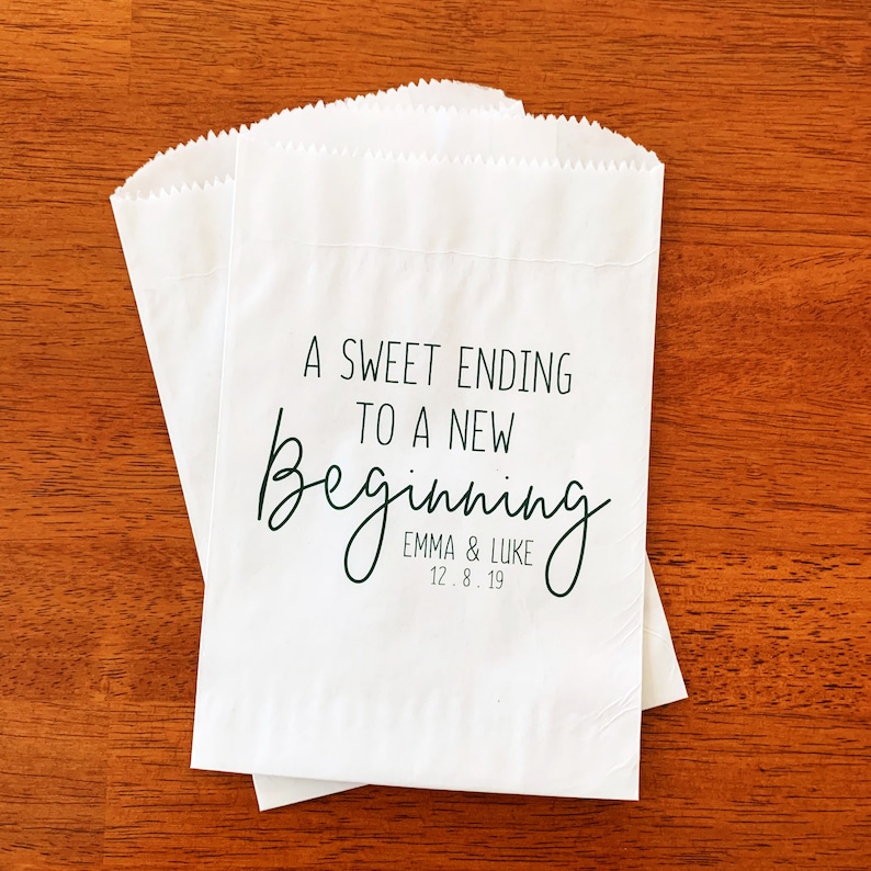 May include: White paper favor bags with a green inscription that reads "A Sweet Ending to a New Beginning" with the names "Emma & Luke" and the date "12.8.19" printed below.